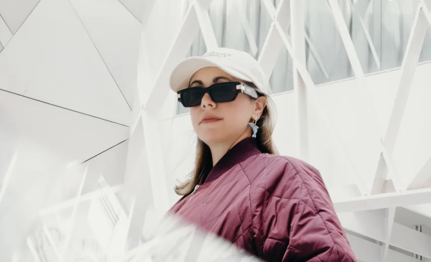 mujer con gorra blanca y gafas de sol negras, pendientes grandes , chaqueta acolchada color granate,