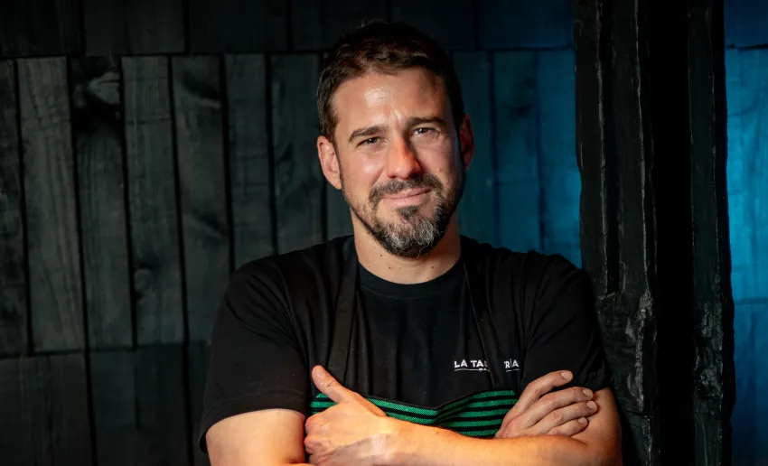 primer plano de hombre con los brazos cruzados, camiseta de manga corta negra, moreno con perilla sonriendo