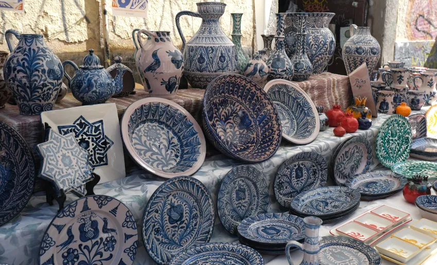 Mesa de mercado llena de piezas de cerámica artesanal —jarras, cuencos y morteros— decoradas con esmaltes verdes, azules y motivos tradicionales, expuestas sobre manteles de tela en un puesto al aire libre.