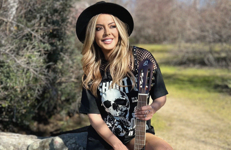 Mujer rubia con sombrero negro y camiseta negra sujetando una guitarra