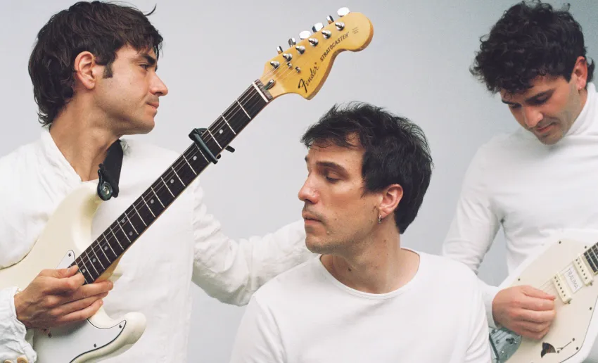 tres chicos vestidos de blanco, fondo neutro blanco, morenos, ninguno mira a cámara, dos de ellos con guitarras en las manos