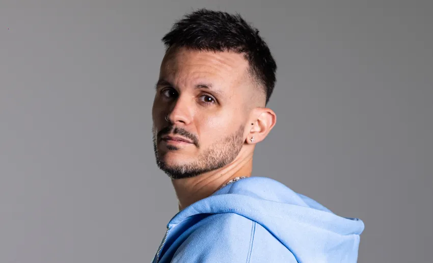 hombre mirando de con el cuerpo de lado, medio de perfil, moreno, leve barba y bigote, sudadera con capucha de algodón en color azul