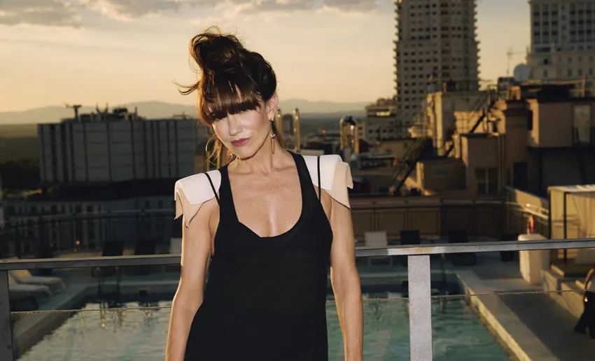 mujer en una terraza con vistas a una ciudad, gran flequillo que le cubre los ojos, pelo recogido, vestido negro de tirantes con grandes hombreras blancas