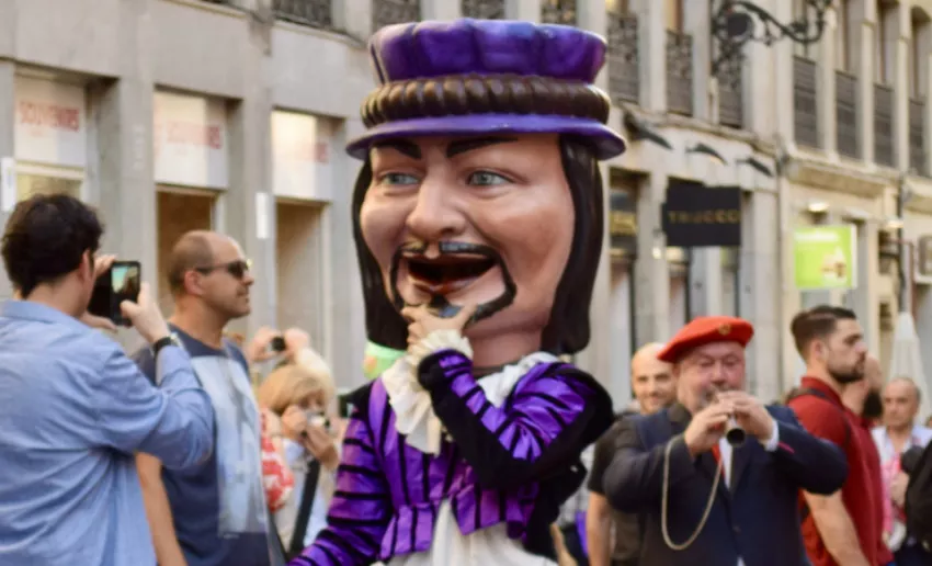 Figura de un gigante festivo con gran cabeza y expresión sonriente, vestido con traje morado y sombrero, desfilando por una calle mientras músicos y varias personas lo acompañan y observan.