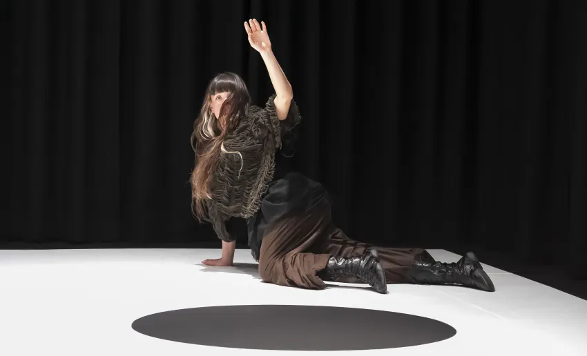 mujer en el suelo vestida de negro, pelo largo que le cubre la cara,  de rodillas y un brazo en alto
