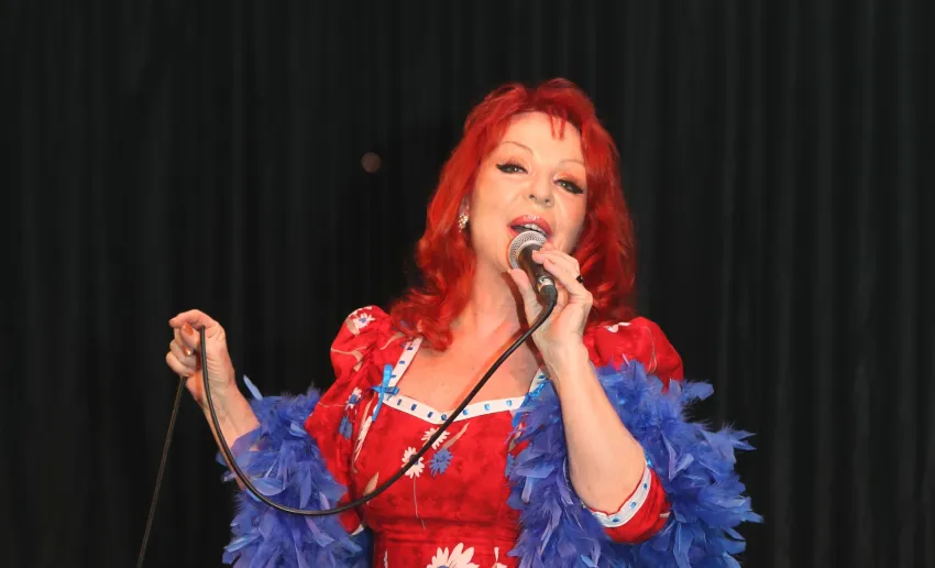 Mujer pelirroja con micrófono en la mano, gesto de cantar, vestido rojo de chulapa y boa azul de plumas, fondo negro